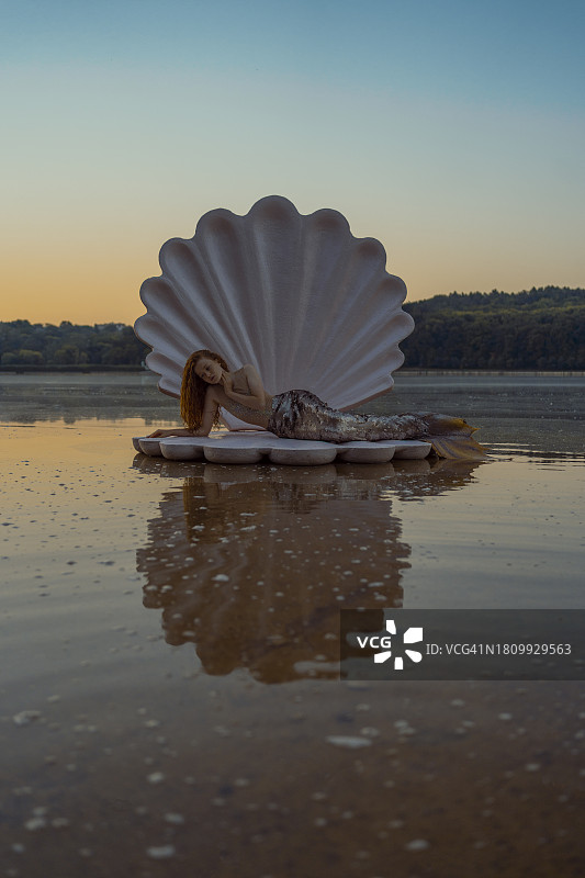 日落时分，海滩上拥有巨型贝壳的美丽红发美人鱼的抽象概念图片素材