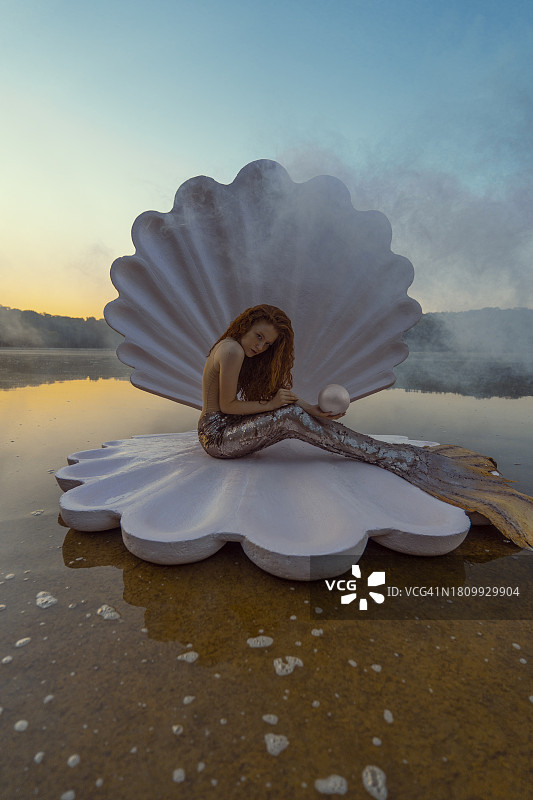 日落时分，海滩上拥有巨型贝壳的美丽红发美人鱼的抽象概念图片素材