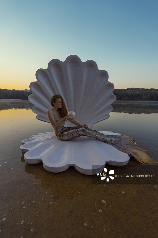 日落时分，海滩上拥有巨型贝壳的美丽红发美人鱼的抽象概念图片素材