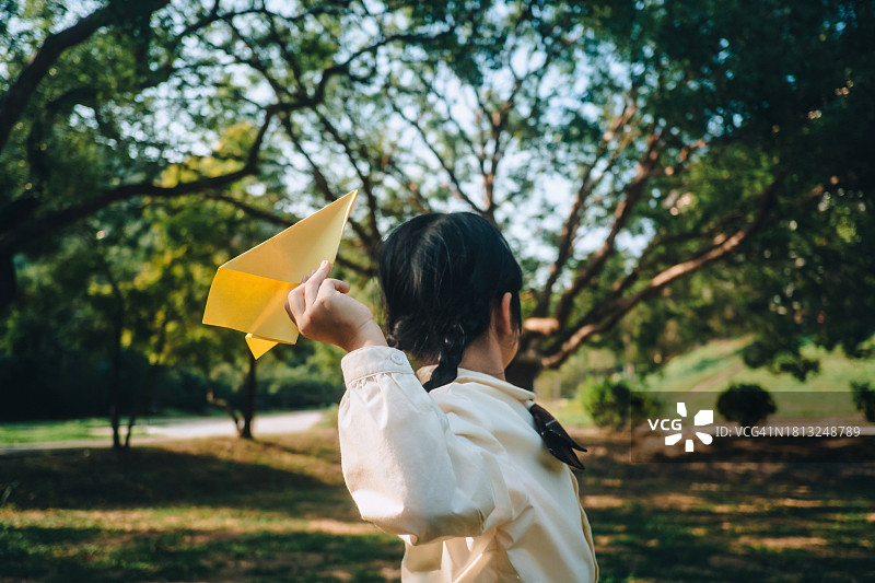 阳光明媚的日子里，在公园里玩纸飞机的年轻亚裔女孩的背影。自由和创造。教育、学生和毕业概念图片素材