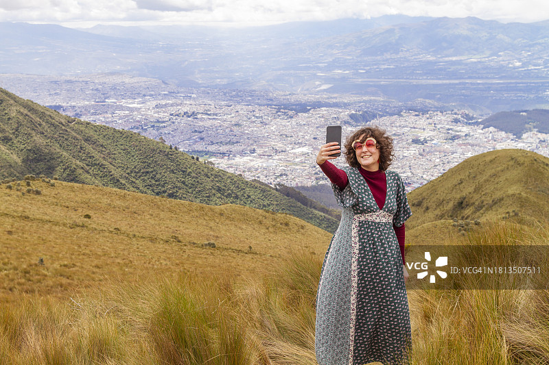 在皮钦查火山顶峰用5G智能手机自拍的年轻女性旅行者肖像，背景是厄瓜多尔首都基多图片素材