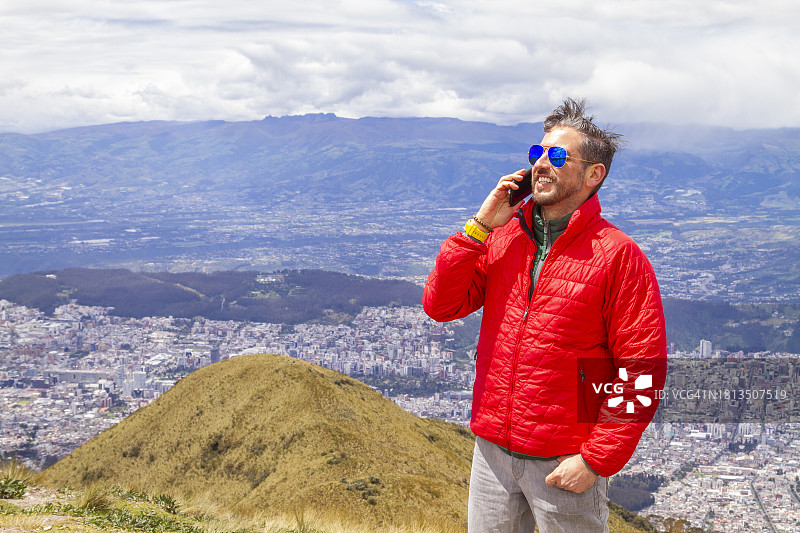 年轻男背包客在钦查火山顶用5G技术通过智能手机交谈，背景为厄瓜多尔首都基多图片素材