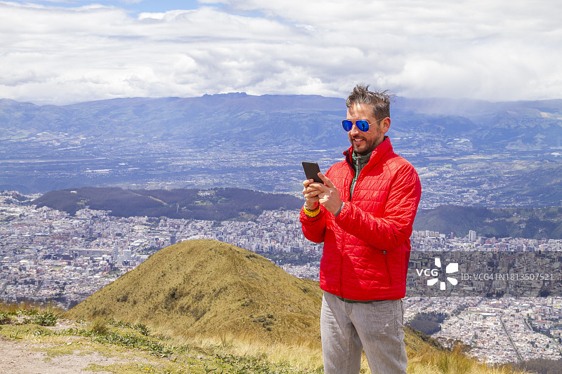 年轻男性独自旅行者在钦查火山顶峰用5G智能手机发送短信，背景是厄瓜多尔首都基多图片素材