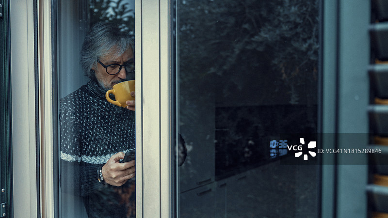 成年男子在窗边查看智能手机，同时喝着热饮图片素材