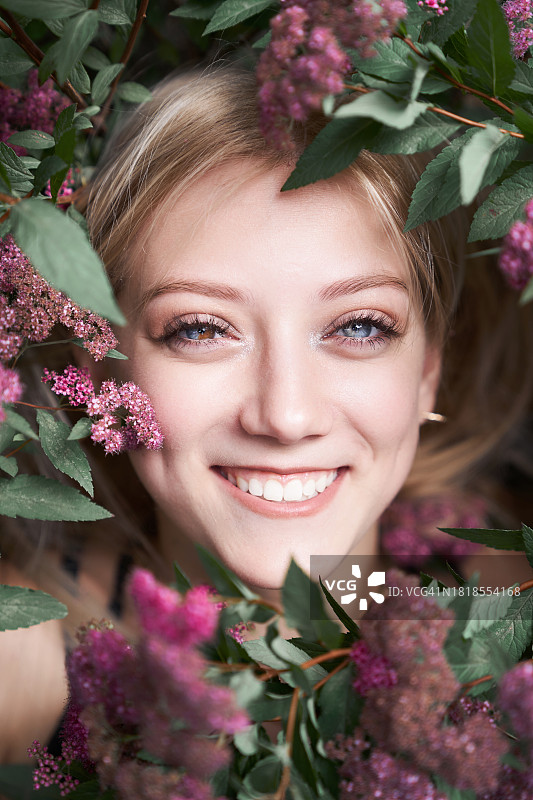 一位眼睛有异色症的年轻少女的美丽特写肖像。金发女人在日本绣线菊丛中。一位年轻快乐的微笑女性的面部特写，脸颊上有酒窝，周围环绕着鲜花。香气和香水概念图片素材