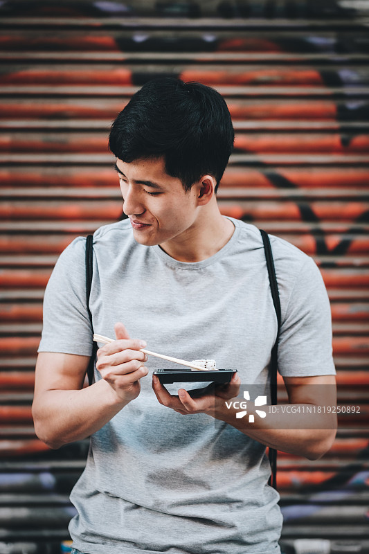 在街上靠着卷帘门吃寿司的年轻微笑的亚洲男子图片素材