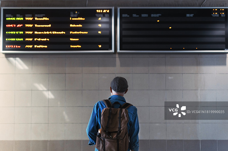 时尚的老年男子在火车站看着火车到站和出发的指示牌，正要回家。老年人在登机前检查列车延误、站台号、出发时间、到达时间。图片素材