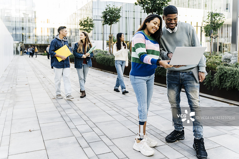 在大学校园外看笔记本电脑的微笑跨种族学生情侣。一位残疾妇女和她的同学正在使用假肢图片素材