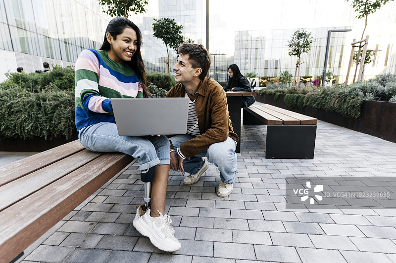 年轻拉丁裔女学生坐长凳上用假肢与同学聊天拿笔记本电脑图片素材