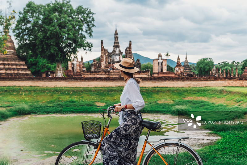 戴着帽子，骑着黄色自行车，在泰国素可泰遗址沉思的年轻女子图片素材