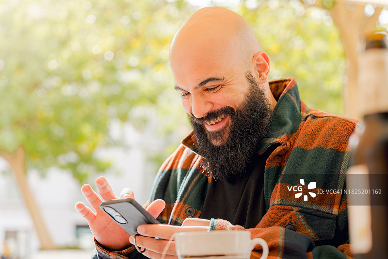 秃顶男人在桌旁使用智能手机微笑，秃顶男人在城市露台上喝茶图片素材