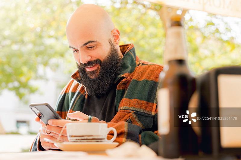秃顶男人在桌旁使用智能手机微笑，秃顶男人在城市露台上喝茶图片素材