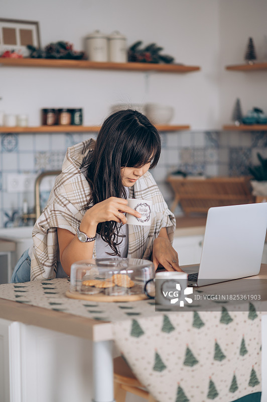 美丽的亚洲女人在厨房喝咖啡，并在笔记本电脑上查看电子邮件。自由职业，远程工作。图片素材