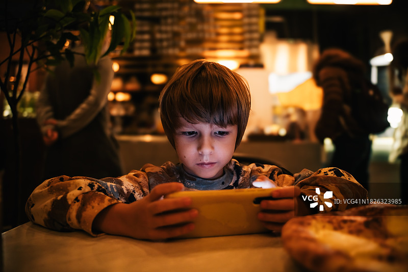 学龄前男孩在黑暗咖啡馆里用手机看视频图片素材