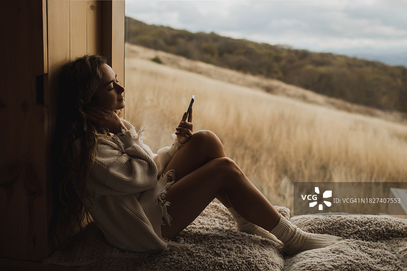 女人在小木屋的窗台上放松，在社交媒体上玩手机图片素材