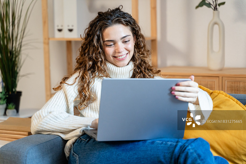 微笑的年轻女人在家里的沙发上使用笔记本电脑图片素材