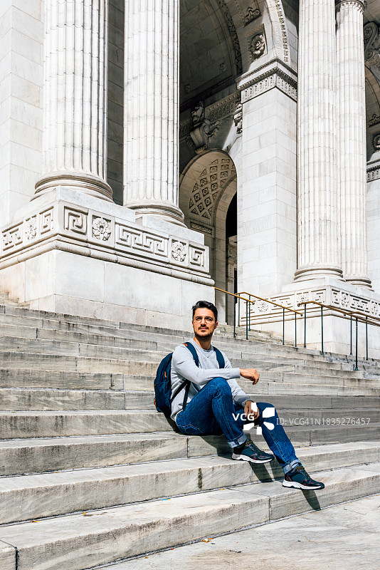 背着背包、戴着眼镜坐在美国纽约公共图书馆台阶上的男人图片素材