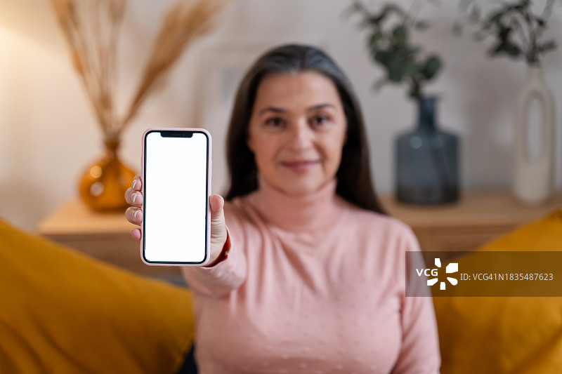 笑容满面的中年妇女在家展示带空白屏幕的智能手机的肖像图片素材