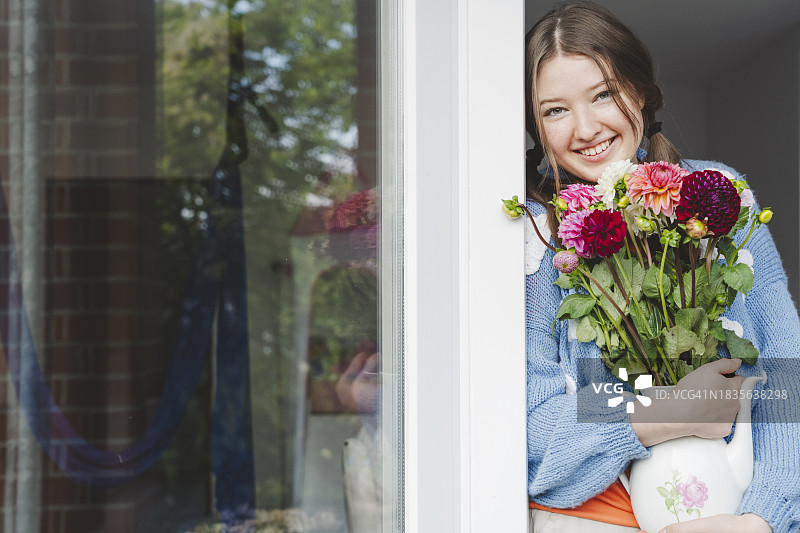 快乐的青少年女孩手捧大丽花，倚靠在窗边图片素材