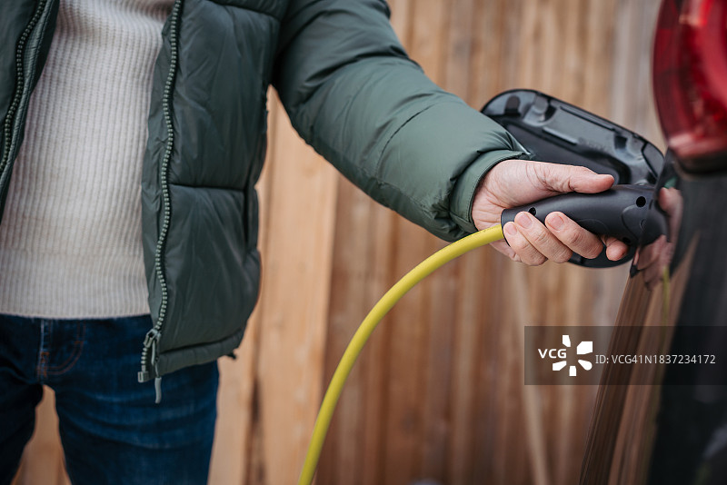 男人在山间小屋前为电动汽车充电，冬季驾驶电动汽车度假图片素材