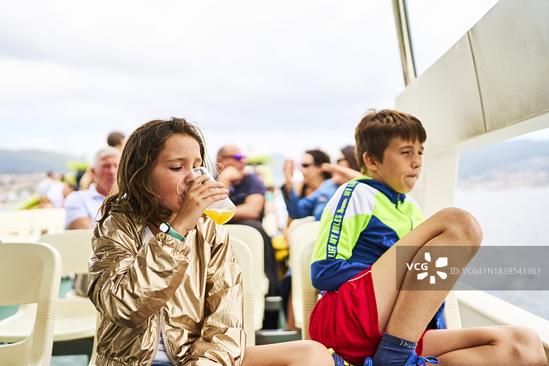 两位游客男孩坐在椅子上，等待时看向远方，女孩在喝果汁，正面照图片素材