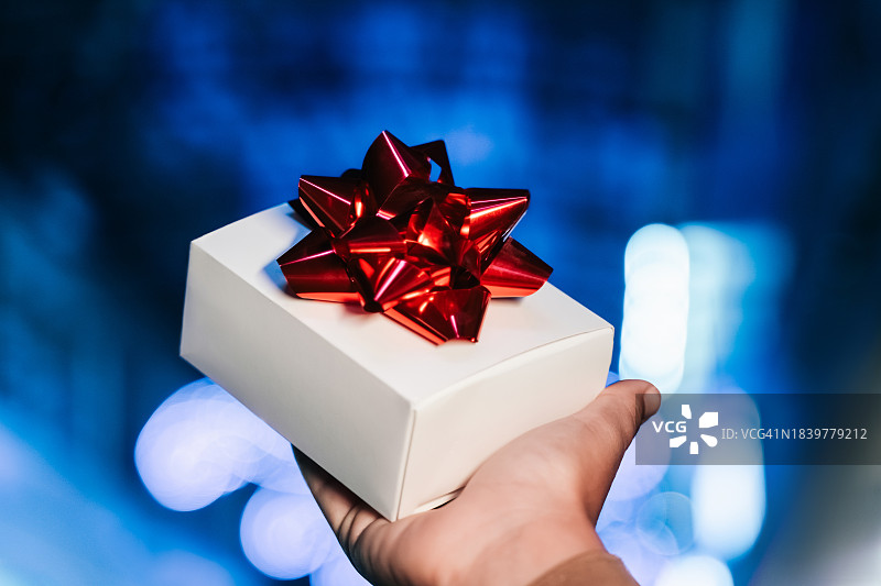 蓝色模糊夜景背景中，手中拿着系着红色蝴蝶结的白色圣诞礼物图片素材
