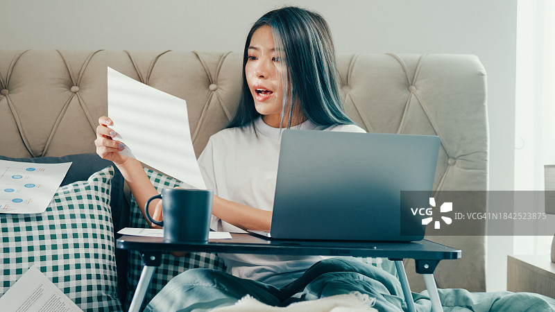 快乐的年轻亚洲女性坐在床上，在折叠笔记本电脑桌上使用电脑和咖啡杯，便携式支架在卧室里拿着文件进行在线视频会议，在家工作图片素材