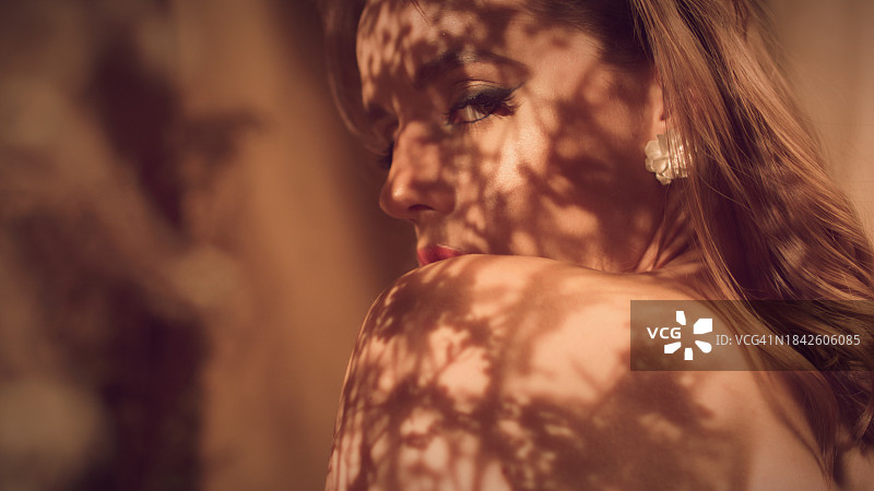 裸露手臂化着妆的性感女人在镜头前卖弄风情，躲在干花后面。抽象花纹阴影图片素材