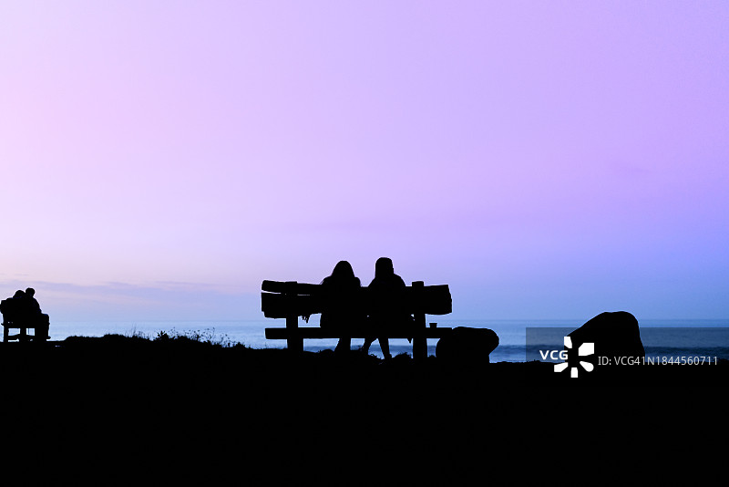两名女子坐在海滩长椅上，眺望日落时海景的背面图图片素材