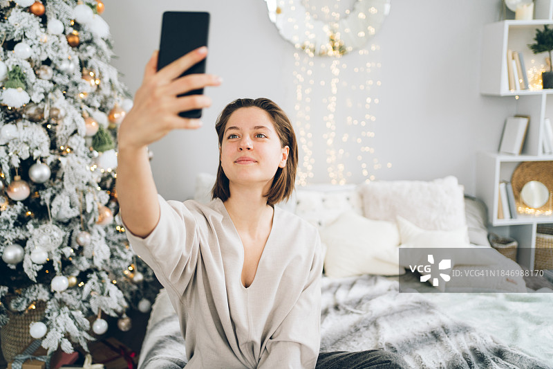 年轻漂亮的女人在装饰着圣诞树的房间里自拍。她坐在一个灯光明亮的房间里的床上。新年庆祝概念。正面视图图片素材