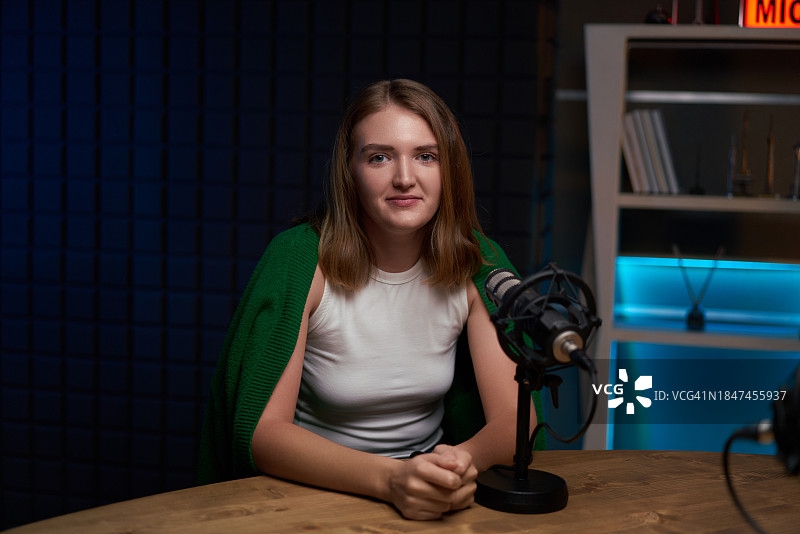快乐的年轻女人在明亮的工作室里，坐在播客桌旁，手持麦克风图片素材