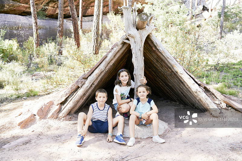 三个孩子坐在干燥的木屋下，在阳光照射下的树木和灌木丛中看着镜头图片素材