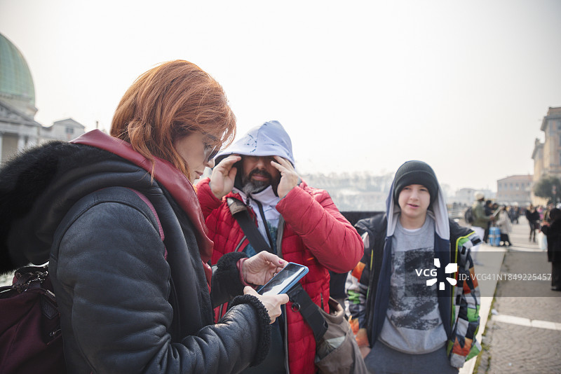中年妇女在使用手机，她的丈夫和儿子在意大利威尼斯等待，一家人图片素材