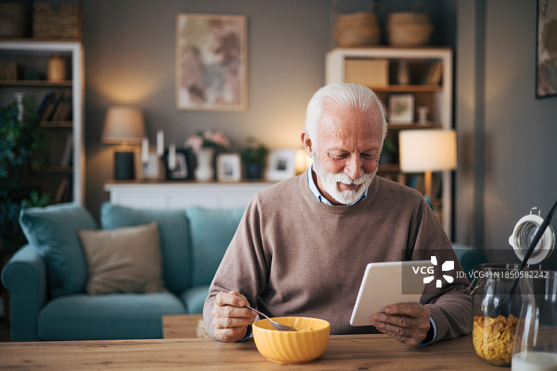 快乐的年长男人在家使用平板电脑工作，并享用一碗麦片作为早餐图片素材