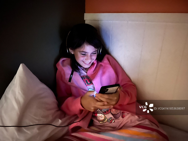 快乐的少女在床上用耳机和智能手机图片素材