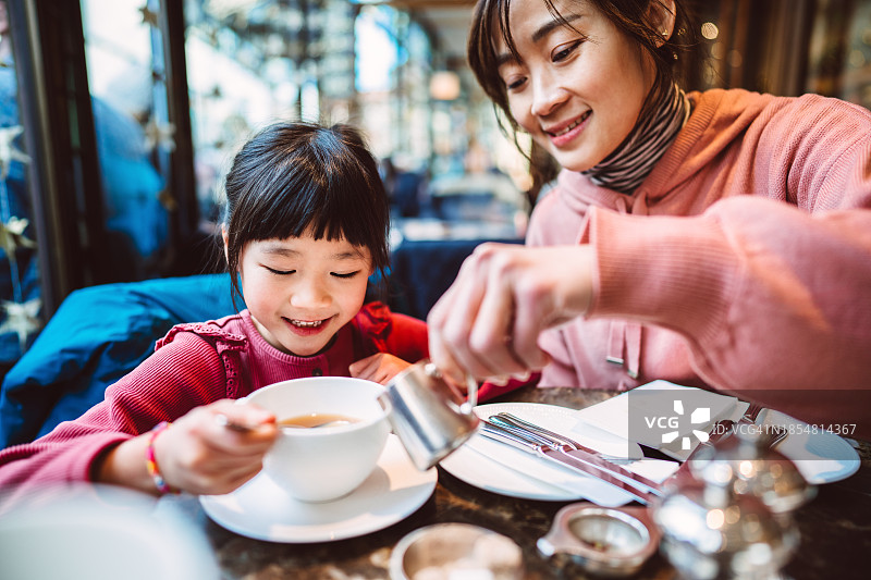 美丽的亚洲妈妈在餐厅与可爱的女儿一起享用下午茶，往热茶杯里倒牛奶图片素材