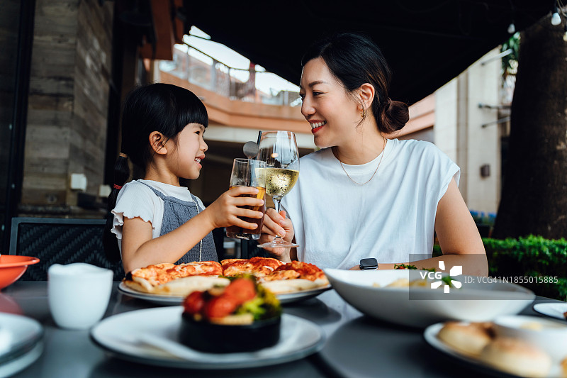 快乐的亚洲家庭在户外餐厅享用美食。年轻的亚洲母亲和女儿在用餐时举杯庆祝，分享母女 bonding 时光。家庭生活方式，一起外出就餐图片素材