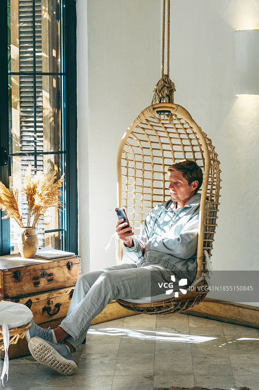 中年男人在悬挂的球形椅子上放松，用手机阅读新闻图片素材