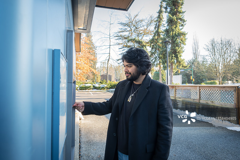 在ATM机上插银行卡的西班牙裔年轻男子，冬日图片素材