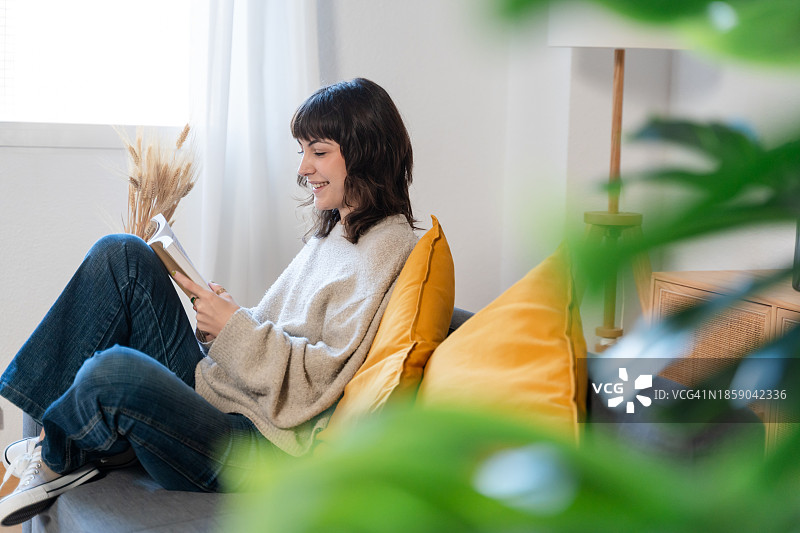 微笑的年轻女人坐在客厅的沙发上看书图片素材