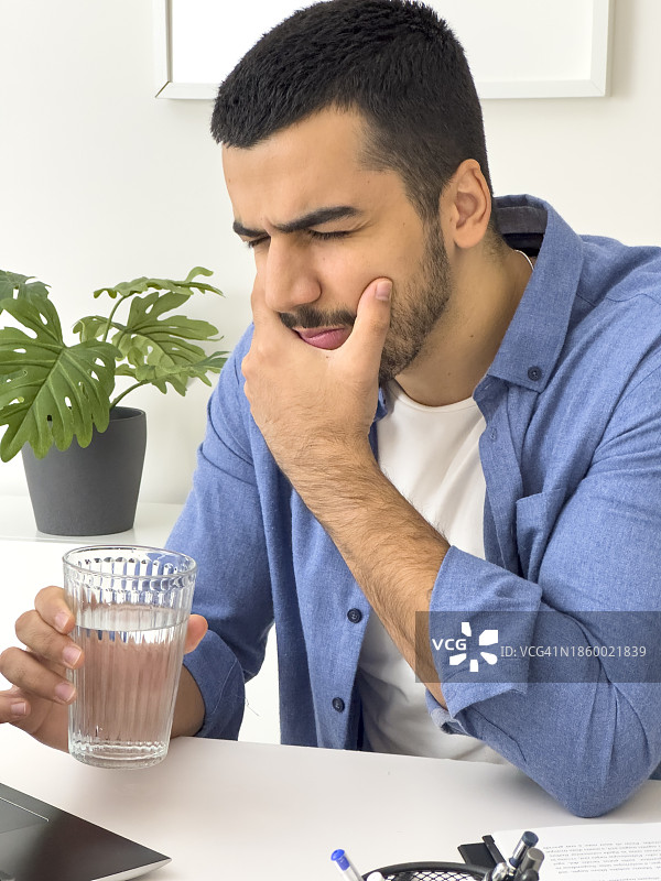喝冷水后牙痛难忍的年轻男人特写。英俊的年轻男人在办公室用电脑时摸着脸颊，因牙齿敏感而感到疼痛图片素材