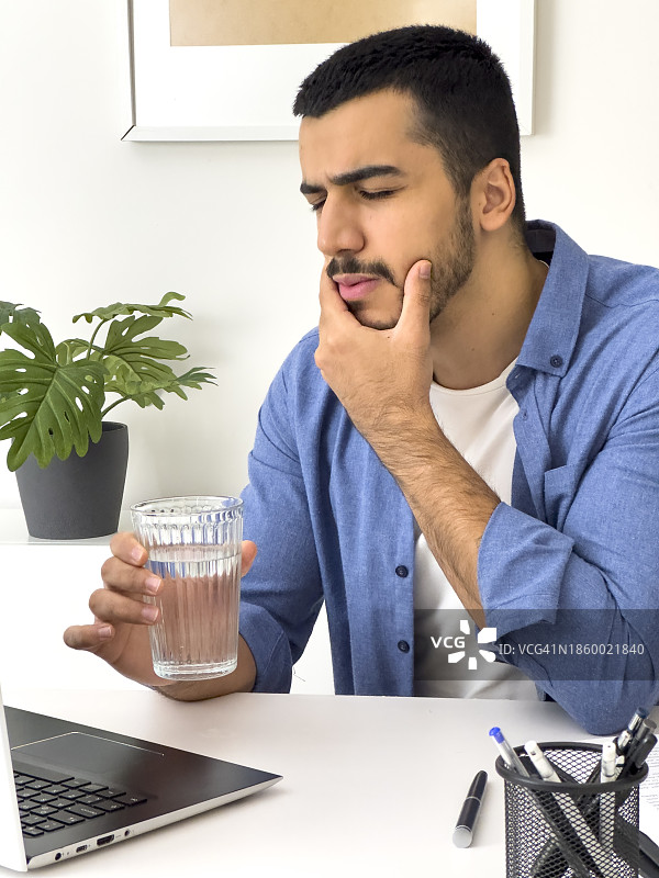 喝冷水后牙痛难忍的年轻男人特写。英俊的年轻男人在办公室用电脑时摸着脸颊，因牙齿敏感而感到疼痛图片素材
