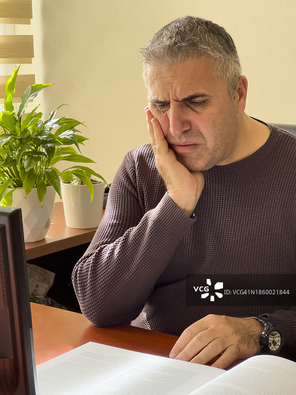 白发商人在办公室用电脑工作，感到严重的牙痛。他触摸脸颊，疼痛并患有敏感性牙痛图片素材