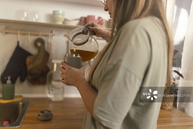 女士将水壶中的热草药茶倒入厨房中的可重复使用杯子中图片素材