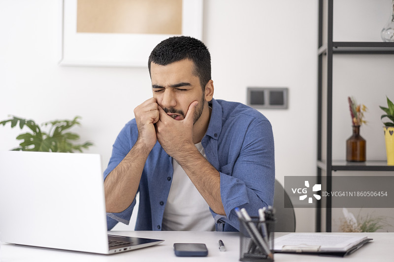 年轻男人牙痛的特写。英俊的年轻男人在家里的办公室用电脑时，摸着脸颊，因牙痛而感到痛苦。图片素材