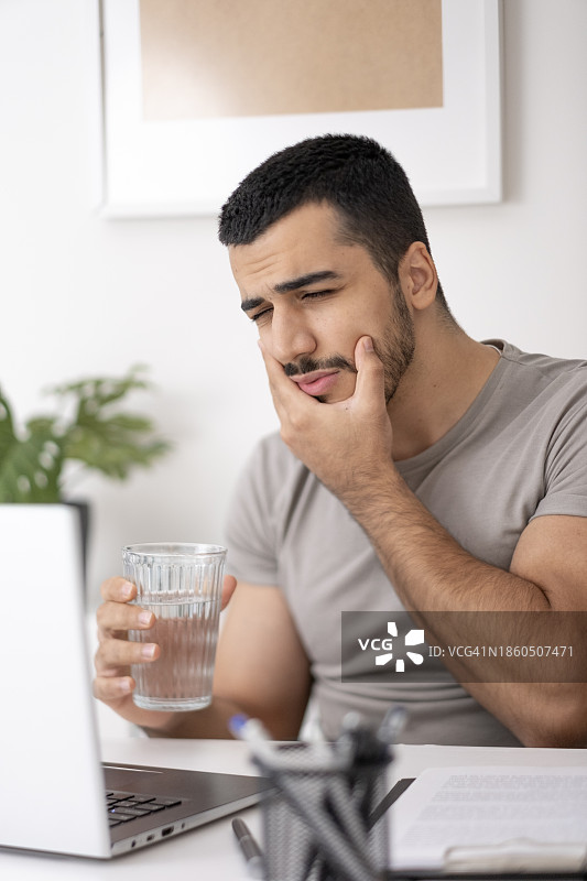 喝冷水后牙痛难忍的年轻男人特写。英俊的年轻男人在办公室用电脑时摸着脸颊，因牙齿敏感而感到疼痛图片素材