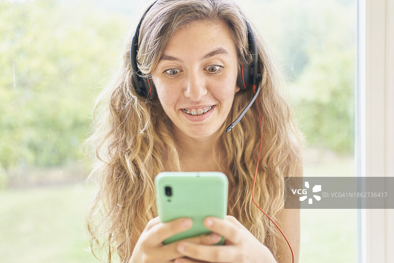 休闲装长发女孩戴耳机持手机，惊讶大笑表情正面照图片素材