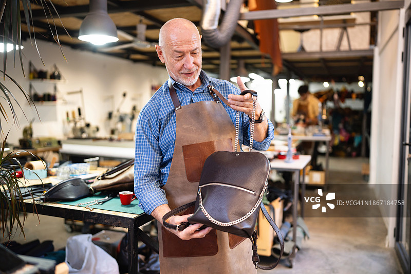 这是他在皮革作坊里制作的一幅白人中年男裁缝拿着皮革背包的肖像图片素材