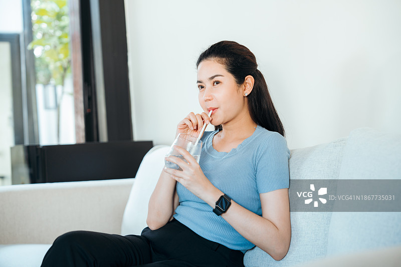 坐在家里客厅沙发上用吸管喝冰水的亚洲女人图片素材