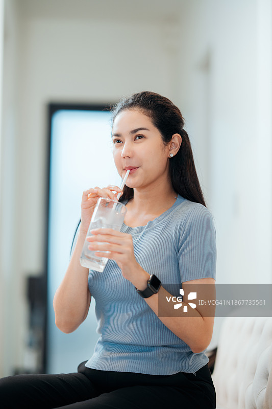 坐在家里客厅沙发上用吸管喝冰水的亚洲女人图片素材